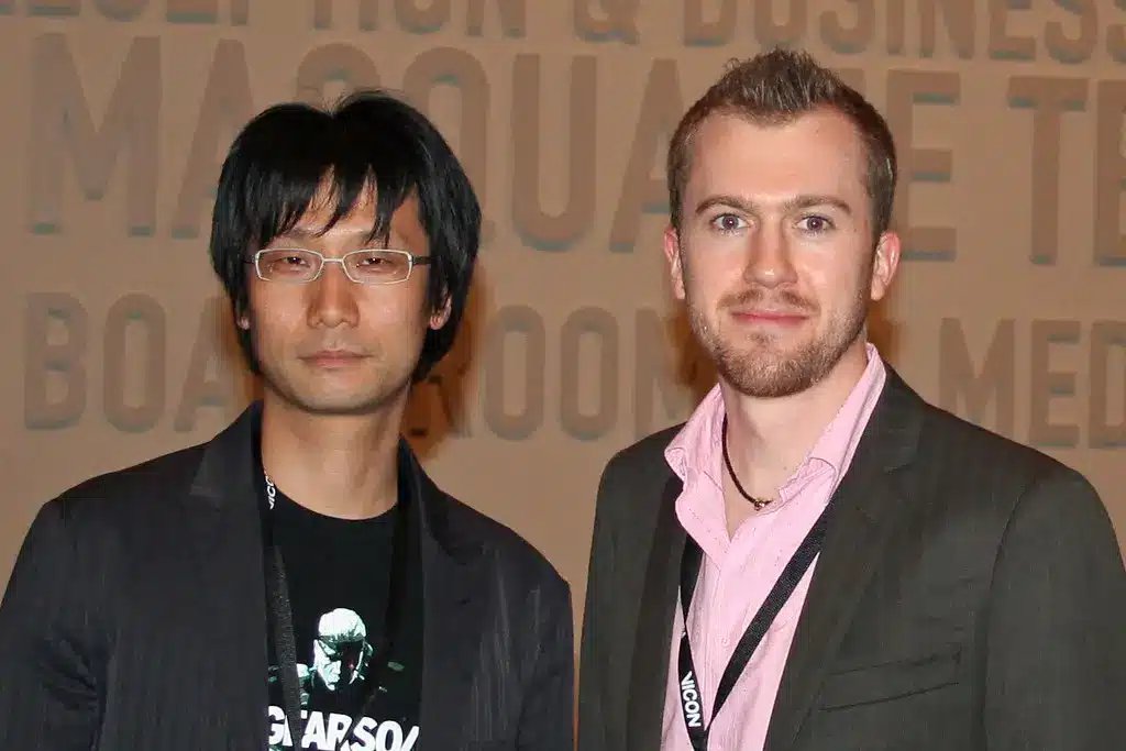 Hideo Kojima usando óculos e um blazer preto, em pé ao lado de outro homem.